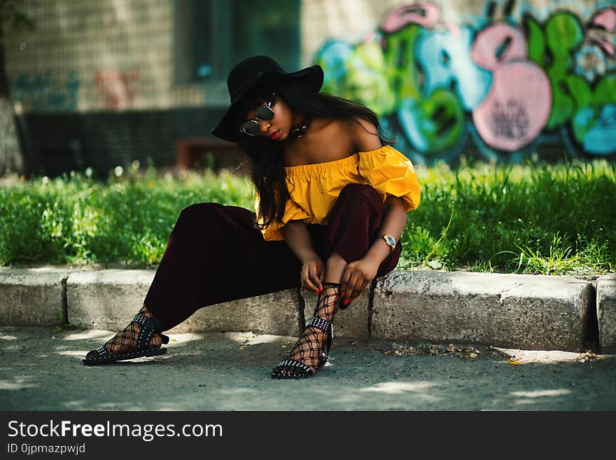 Woman Wearing Yellow Off-shoulder Top and Black Pants Sitting on Sidewalk Fixing Lace Sandals