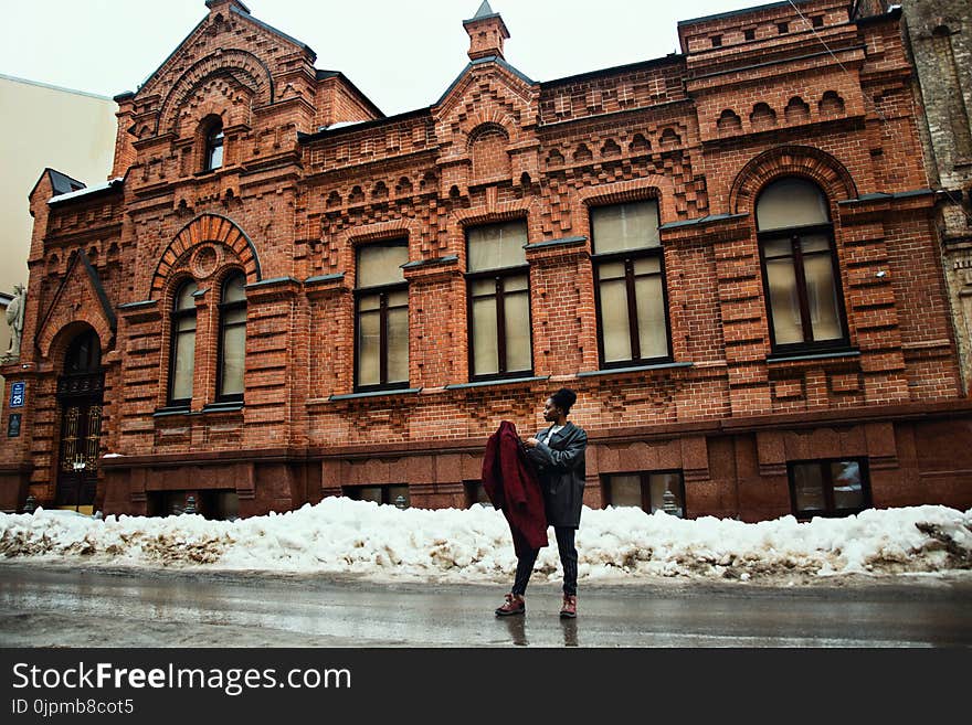 Man Holding Red Coat Standing on Street Near Brown Building