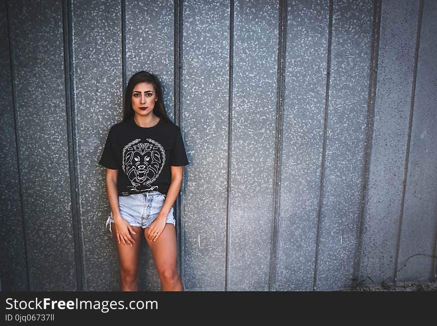 Woman Wearing Black Lion Graphic Crew-neck Shirt and Gray Denim Short Shorts Leaning on Gray Wall