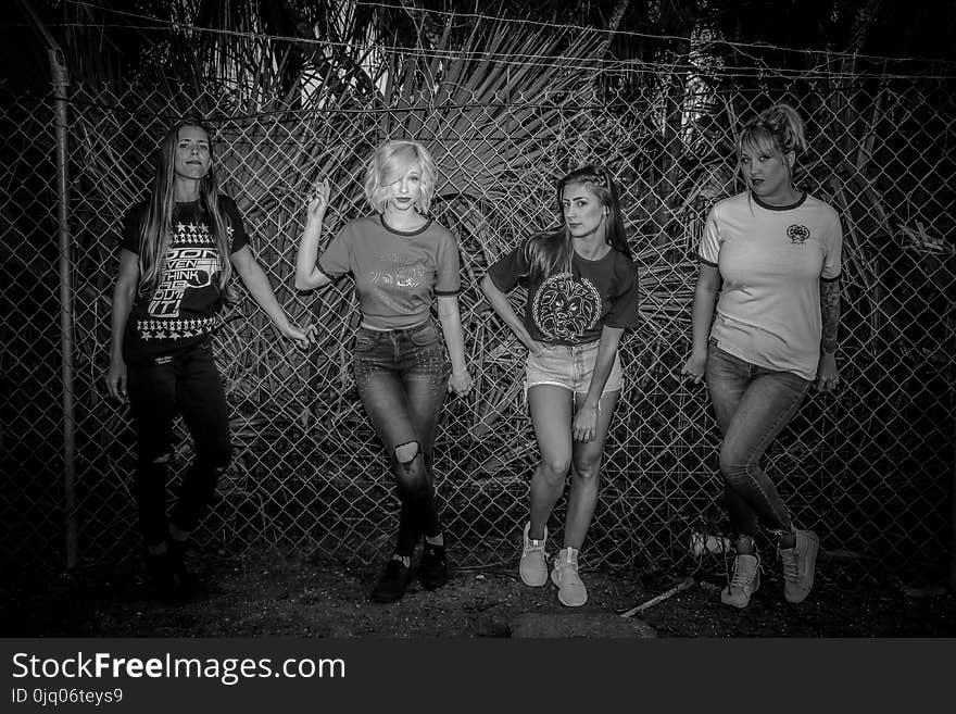 Grayscale Photo of Four Woman Standing Near on Link Fence