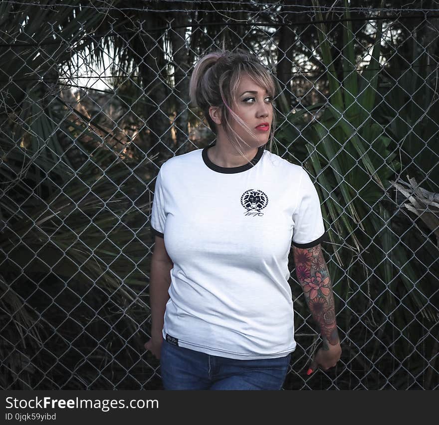 Woman Wearing White Crew-neck T-shirt With Black Trim and Blue Denim Bottoms Beside Wire Fence