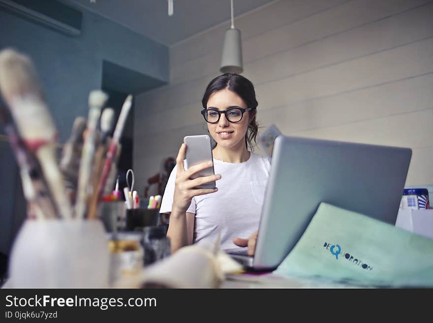 Woman in White T-shirt Holding Smartphone in Front of Laptop