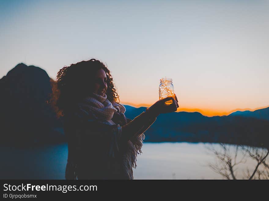 Woman Holding Mason Jar With String Light With Lake and Mountain over View during Golden Hour