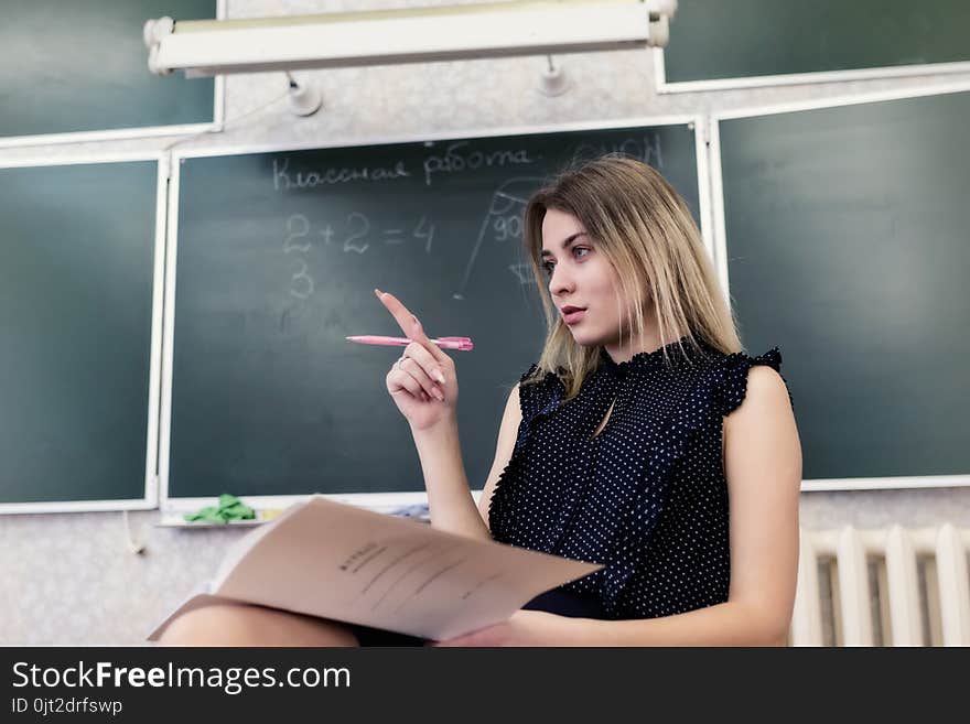 Strict young blonde teacher sits on a chair in the classroom against a blackboard and reflects on who to check homework. Strict young blonde teacher sits on a chair in the classroom against a blackboard and reflects on who to check homework