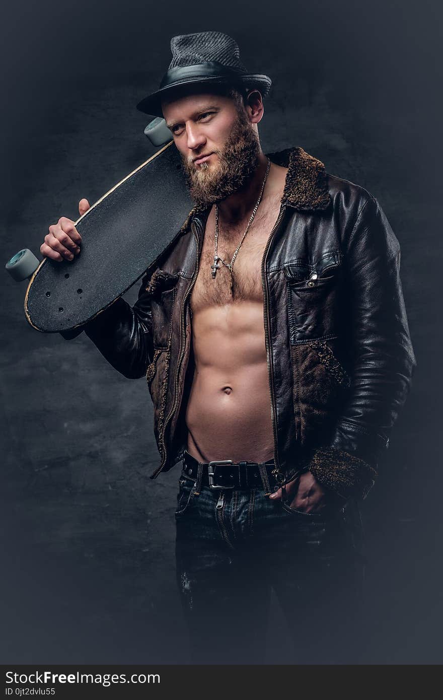 Bearded male dressed in leather jackett holds skateboard.