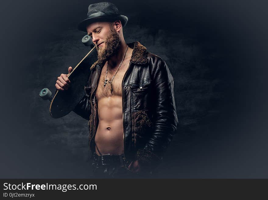 Bearded male dressed in leather jackett holds skateboard.