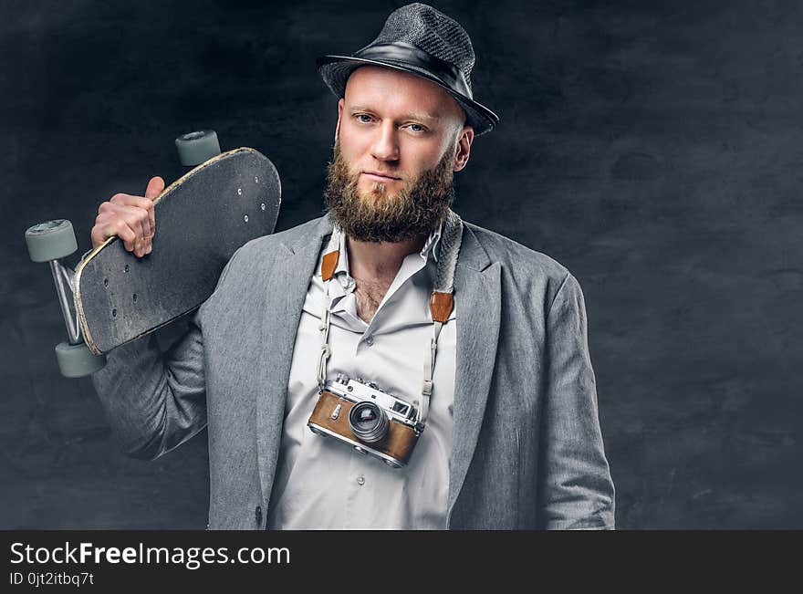 Bearded male in a suit and holds skateboard and photo camera.