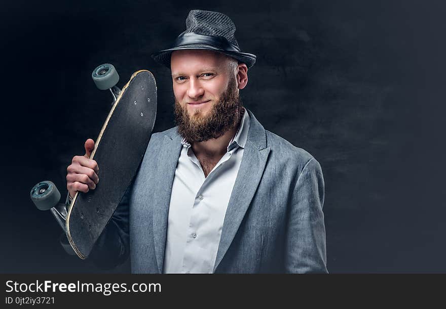 Bearded male in a suit and felt hat holds skateboard.