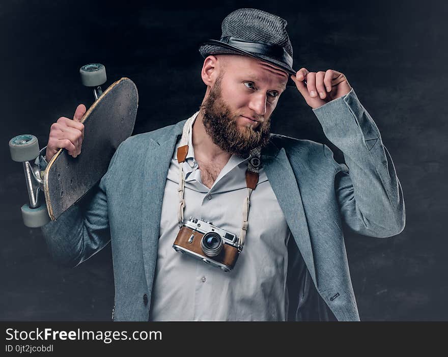 Stylish bearded male in a suit and felt hat holds skateboard and the vintage SLR photo camera. Stylish bearded male in a suit and felt hat holds skateboard and the vintage SLR photo camera.