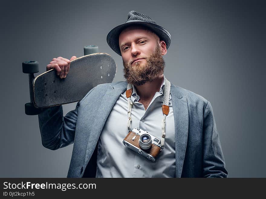 Bearded man in a suit holds skateboard and SLR photo camera.