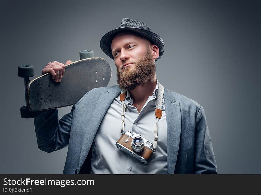 Bearded man in a suit holds skateboard and SLR photo camera.