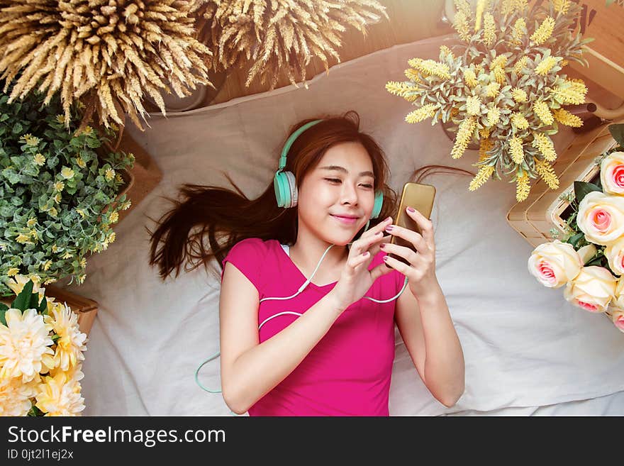 Happy Young Woman Laying On The Floor To Listening Music Via Smart Phone. Girl Relaxing In Cozy Place, Surrounded By Flower, Top