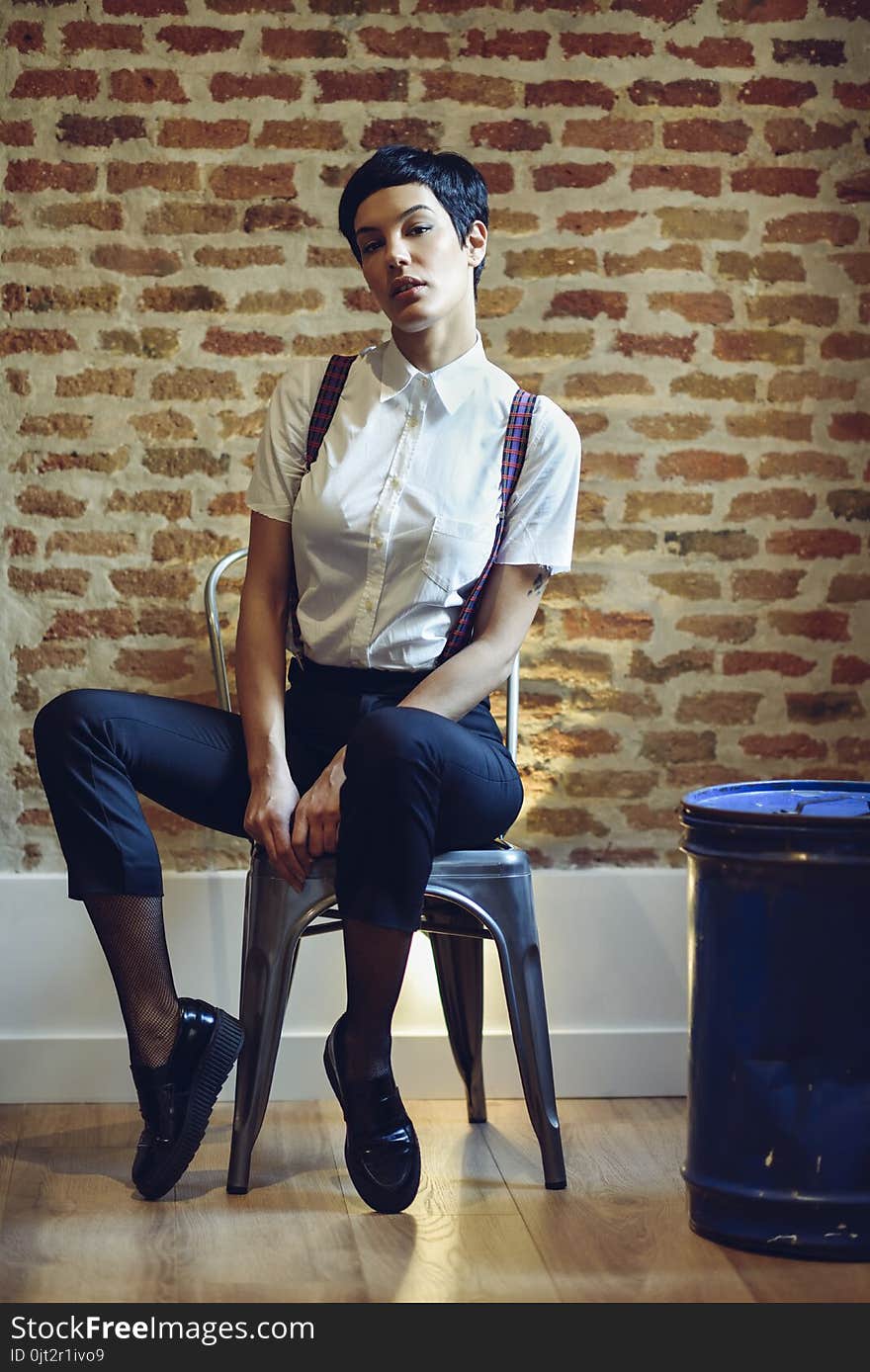 Beautiful young woman, model of fashion, with very short haircut, looking at camera sitting on chair, with a brick wall in the background. Beauty and hairstyles concept.