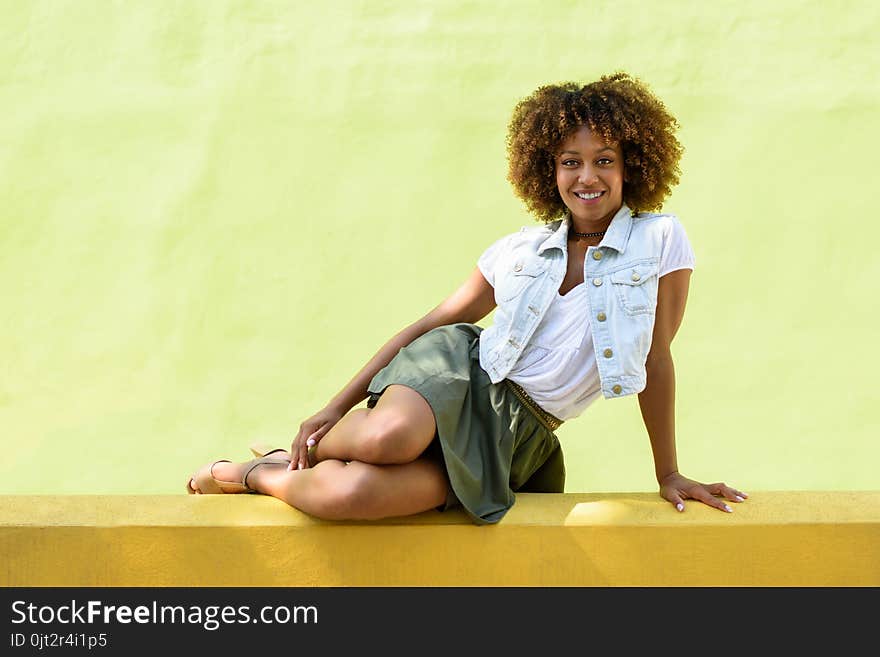 Young black woman, afro hairstyle, sitting on a wall smiling. Girl, model of fashion, wearing casual clothes in urban background. Female with skirt, denim vest and high heels. Young black woman, afro hairstyle, sitting on a wall smiling. Girl, model of fashion, wearing casual clothes in urban background. Female with skirt, denim vest and high heels.