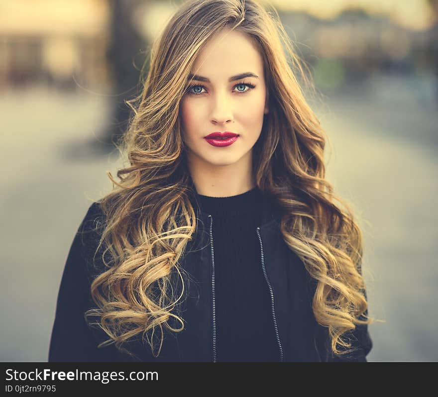 Close-up portrait of young blonde girl with beautiful blue eyes wearing black jacket outdoors. Pretty russian female with long wavy hair hairstyle. Woman in urban background. Close-up portrait of young blonde girl with beautiful blue eyes wearing black jacket outdoors. Pretty russian female with long wavy hair hairstyle. Woman in urban background.