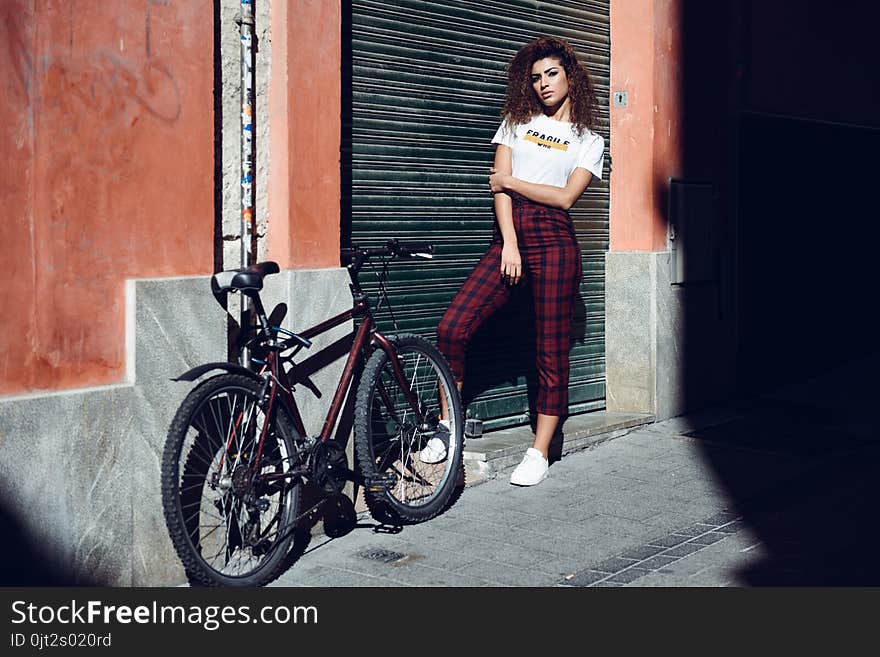 Beautiful young arabic woman with black curly hairstyle. Arab girl in casual clothes in the street near a bicycle. Girl power concept. Beautiful young arabic woman with black curly hairstyle. Arab girl in casual clothes in the street near a bicycle. Girl power concept.