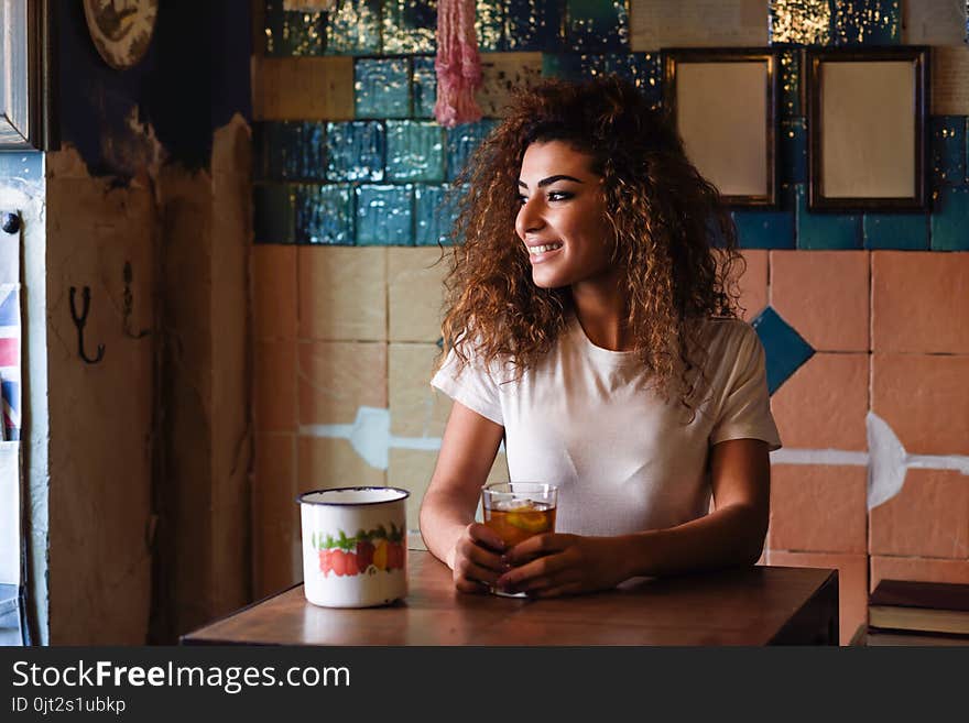 Young arabic smiling woman with black curly hairstyle sitting in a beautiful bar with vintage decoration. Arab girl in casual clothes drinking a soda. Young arabic smiling woman with black curly hairstyle sitting in a beautiful bar with vintage decoration. Arab girl in casual clothes drinking a soda.