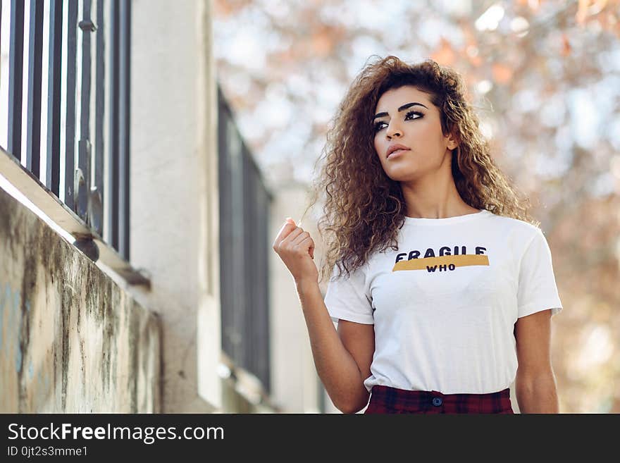 Beautiful young arabic woman with black curly hairstyle. Arab girl in casual clothes in the street. Happy female wearing white t-shirt and checked pants.