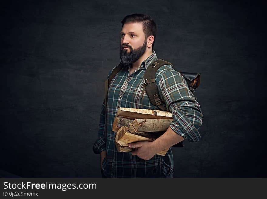 Bearded middle age male holds backpack and firewoods over dark grey background. Bearded middle age male holds backpack and firewoods over dark grey background.