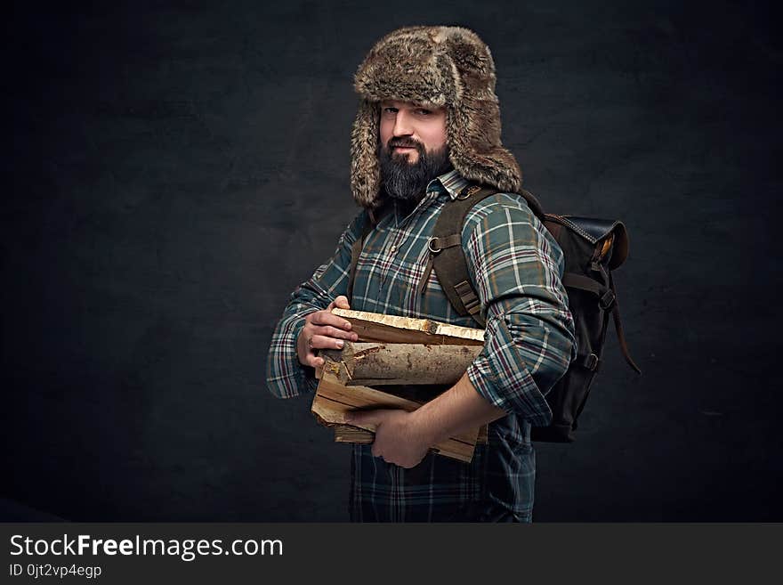 Bearded middle age male in a fur hat holds backpack and firewoods over dark grey background. Bearded middle age male in a fur hat holds backpack and firewoods over dark grey background.