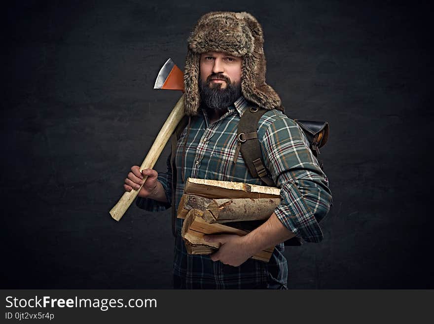 Brutal bearded middle age male dressed in a plaid shirt and a fur hat holds an axe and firewoods over dark grey background. Brutal bearded middle age male dressed in a plaid shirt and a fur hat holds an axe and firewoods over dark grey background.