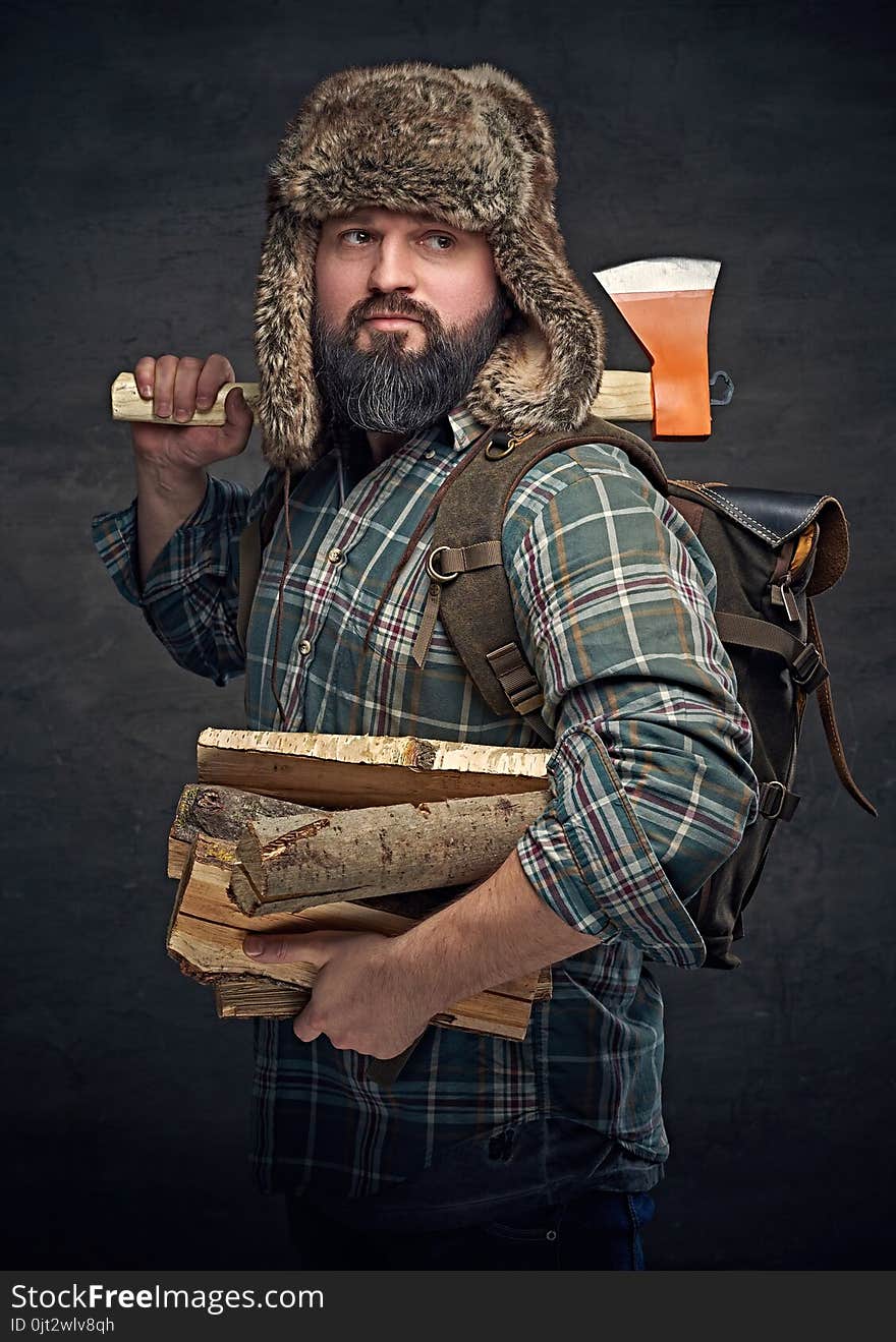 Brutal bearded middle age male dressed in a plaid shirt and a fur hat holds an axe and firewoods over dark grey background. Brutal bearded middle age male dressed in a plaid shirt and a fur hat holds an axe and firewoods over dark grey background.