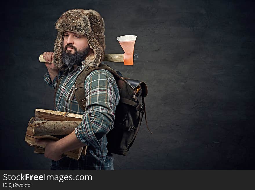 Brutal bearded middle age male dressed in a plaid shirt and a fur hat holds an axe and firewoods over dark grey background. Brutal bearded middle age male dressed in a plaid shirt and a fur hat holds an axe and firewoods over dark grey background.