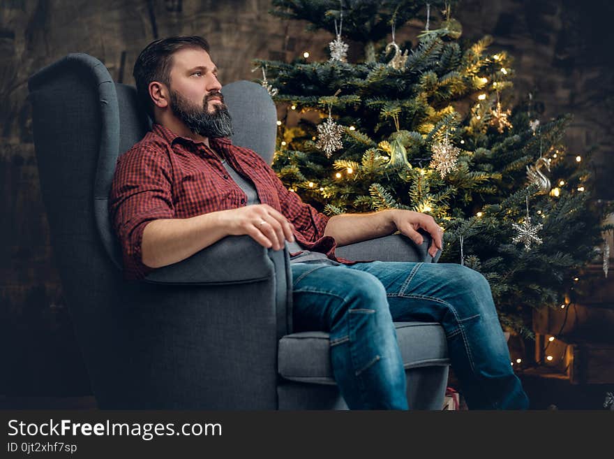 A bearded middle age male dressed in a plaid shirt and jeans sits on a chair over Christmas illumination and fir tree in background. A bearded middle age male dressed in a plaid shirt and jeans sits on a chair over Christmas illumination and fir tree in background.