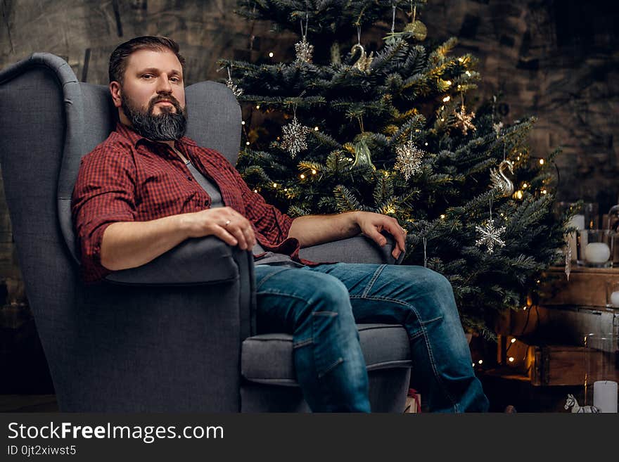 A bearded middle age male dressed in a plaid shirt and jeans sits on a chair over Christmas illumination and fir tree in background. A bearded middle age male dressed in a plaid shirt and jeans sits on a chair over Christmas illumination and fir tree in background.