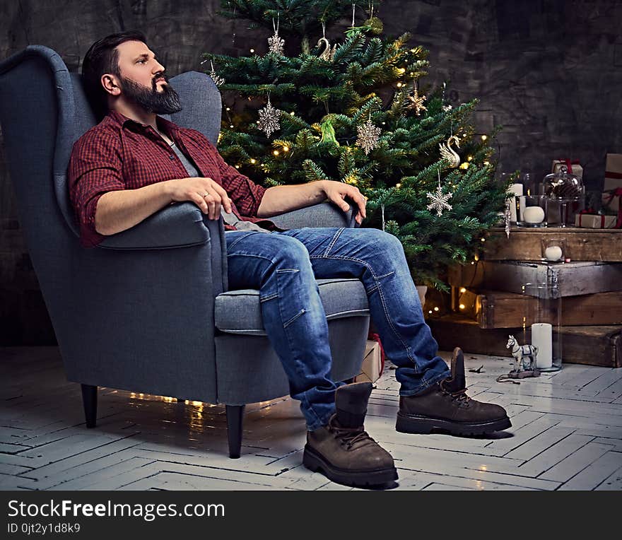 A bearded middle age male dressed in a plaid shirt and jeans sits on a chair over Christmas illumination and fir tree in background. A bearded middle age male dressed in a plaid shirt and jeans sits on a chair over Christmas illumination and fir tree in background.