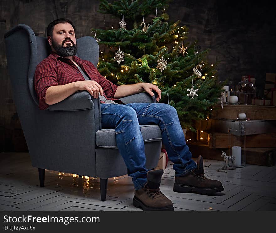 A bearded middle age male dressed in a plaid shirt and jeans sits on a chair over Christmas illumination and fir tree in background. A bearded middle age male dressed in a plaid shirt and jeans sits on a chair over Christmas illumination and fir tree in background.