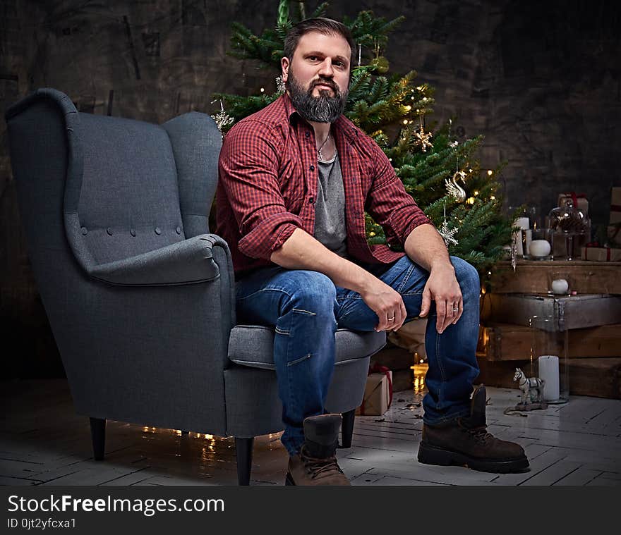 A bearded middle age male dressed in a plaid shirt and jeans sits on a chair over Christmas illumination and fir tree in background. A bearded middle age male dressed in a plaid shirt and jeans sits on a chair over Christmas illumination and fir tree in background.