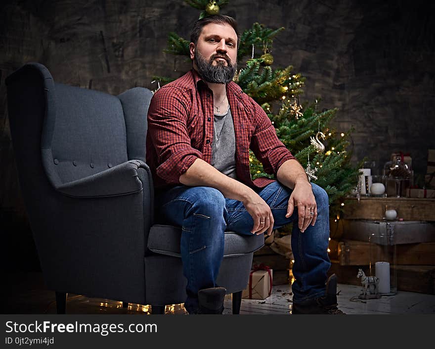 A bearded middle age male dressed in a plaid shirt and jeans sits on a chair over Christmas illumination and fir tree in background. A bearded middle age male dressed in a plaid shirt and jeans sits on a chair over Christmas illumination and fir tree in background.