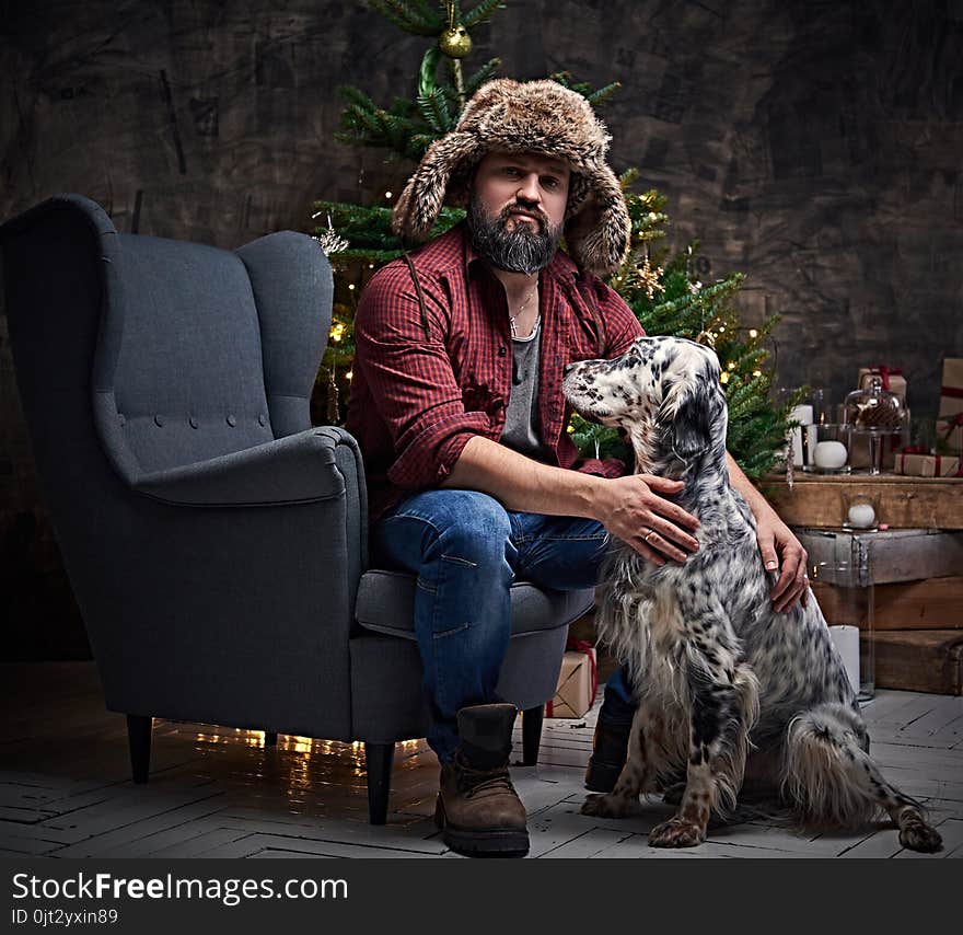 Bearded middle age male dressed in a plaid shirt and fur hat and Irish setter with fir tree and Christmas decoration on background. Bearded middle age male dressed in a plaid shirt and fur hat and Irish setter with fir tree and Christmas decoration on background.