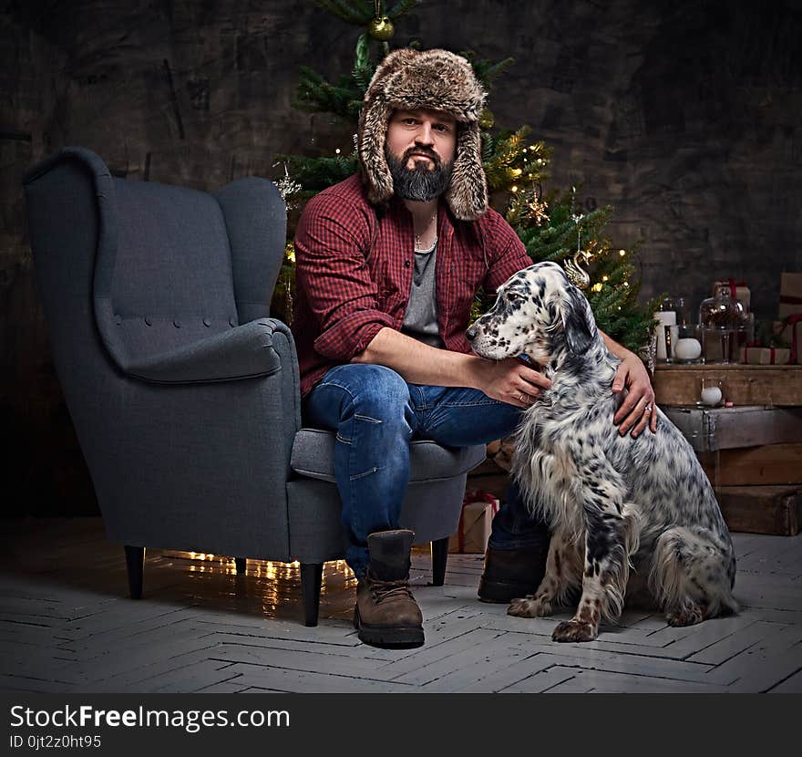 Bearded middle age male dressed in a plaid shirt and fur hat and two Irish setter dogs with fir tree and Christmas decoration on background. Bearded middle age male dressed in a plaid shirt and fur hat and two Irish setter dogs with fir tree and Christmas decoration on background.