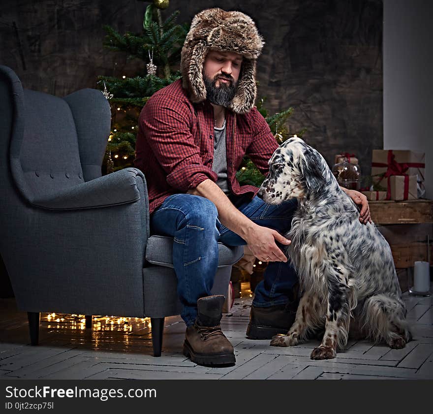 Bearded middle age male dressed in a plaid shirt and fur hat and two Irish setter dogs with fir tree and Christmas decoration on background. Bearded middle age male dressed in a plaid shirt and fur hat and two Irish setter dogs with fir tree and Christmas decoration on background.