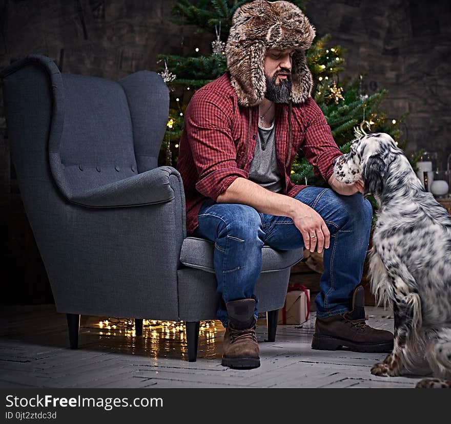 Bearded middle age male dressed in a plaid shirt and fur hat and two Irish setter dogs with fir tree and Christmas decoration on background. Bearded middle age male dressed in a plaid shirt and fur hat and two Irish setter dogs with fir tree and Christmas decoration on background.