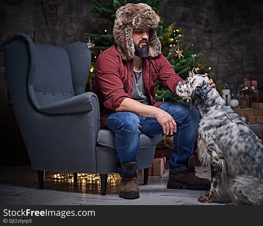 Bearded middle age male dressed in a plaid shirt and fur hat and two Irish setter dogs with fir tree and Christmas decoration on background. Bearded middle age male dressed in a plaid shirt and fur hat and two Irish setter dogs with fir tree and Christmas decoration on background.