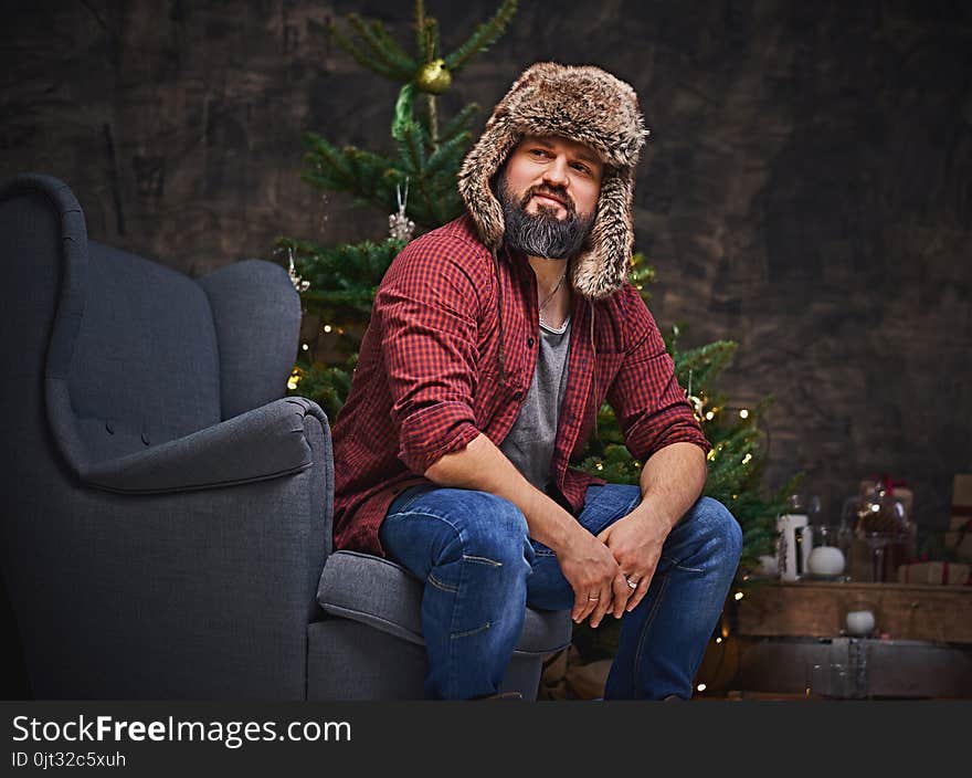 Bearded middle age male dressed in plaid shirt, jeans and fur hat sits on a chair over Christmas illumination and fir tree on background. Bearded middle age male dressed in plaid shirt, jeans and fur hat sits on a chair over Christmas illumination and fir tree on background.