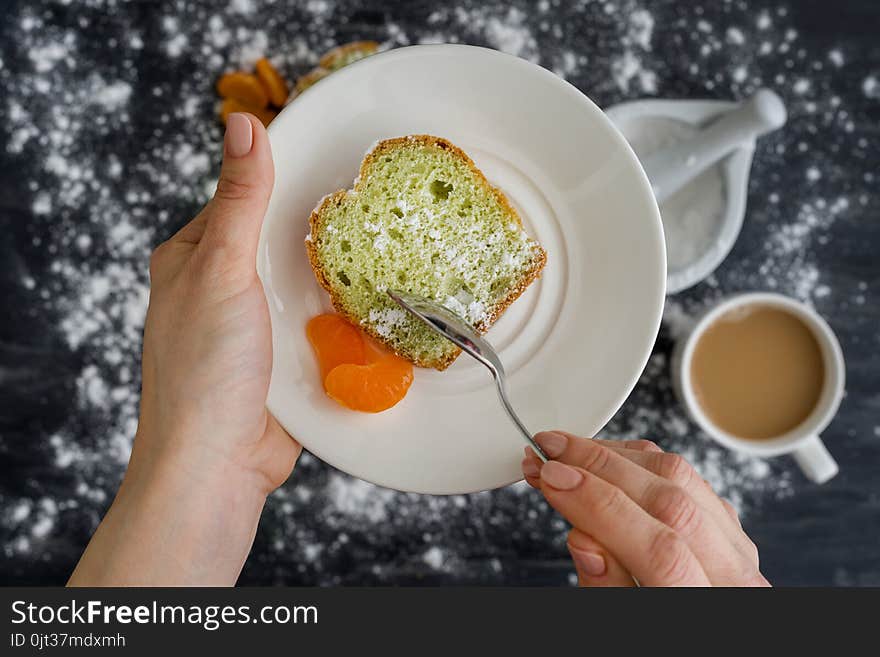 Mint Cake On A Plate With Tangerines In Hands. The Background Is A Dark Surface Sprinkled With Powdered Sugar, A Cup Of Coffee.