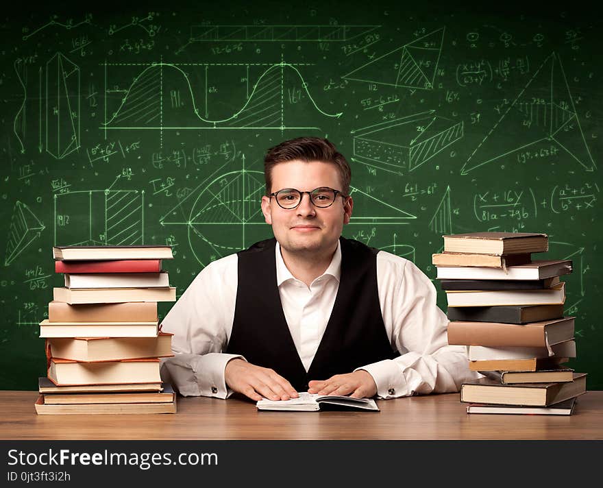 A young passionate male teacher sitting at school desk, reading a book, with area algorythm calculations and numbers on the blackboard concept. A young passionate male teacher sitting at school desk, reading a book, with area algorythm calculations and numbers on the blackboard concept.