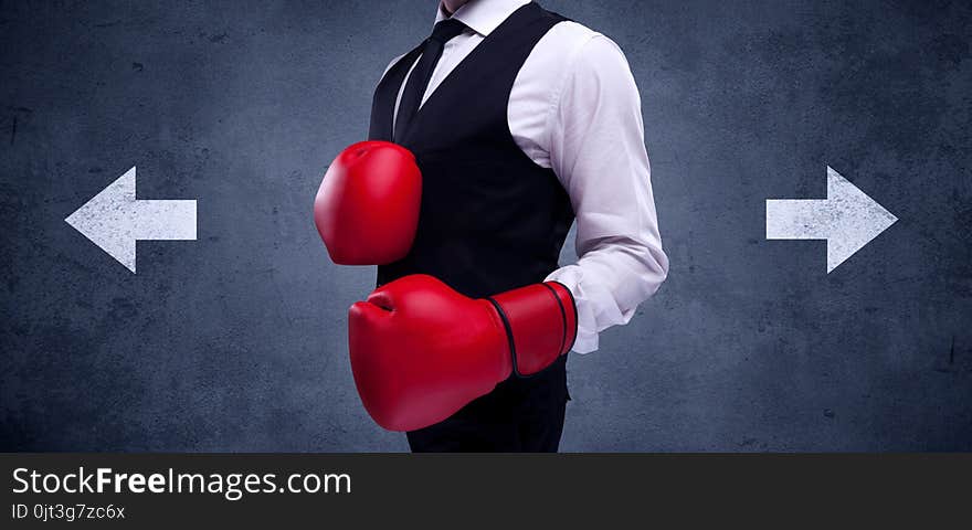 A confident businessman standing with red boxing gloves on his hand in front of arrows pointing in different directions on urban wall background concept. A confident businessman standing with red boxing gloves on his hand in front of arrows pointing in different directions on urban wall background concept.