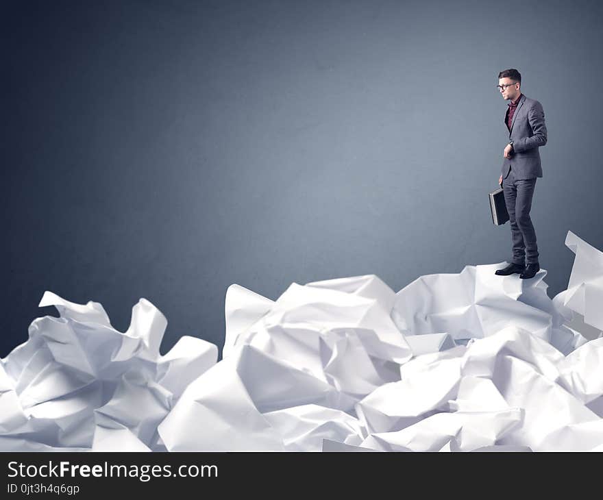 Thoughtful young businessman standing on a pile of crumpled paper with a blueish grey background. Thoughtful young businessman standing on a pile of crumpled paper with a blueish grey background