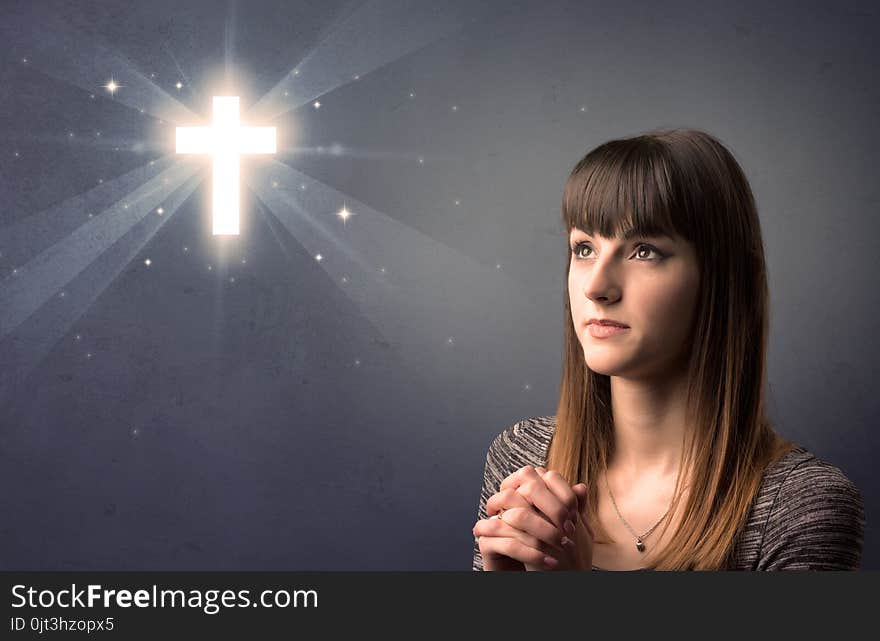 Young woman praying on a grey background with a shiny cross above her. Young woman praying on a grey background with a shiny cross above her