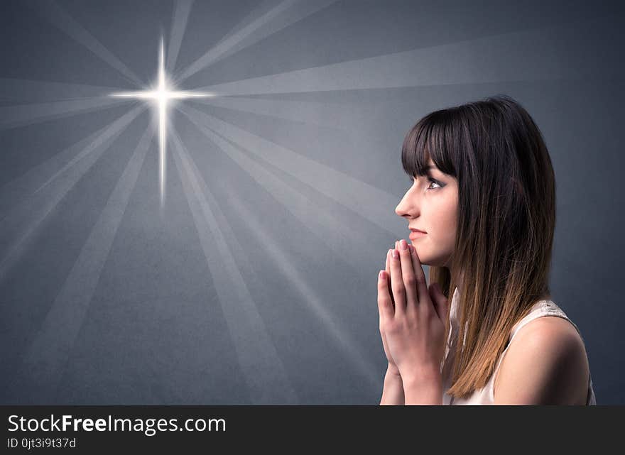 Young woman praying on a grey background with a shiny cross silhouette above her. Young woman praying on a grey background with a shiny cross silhouette above her