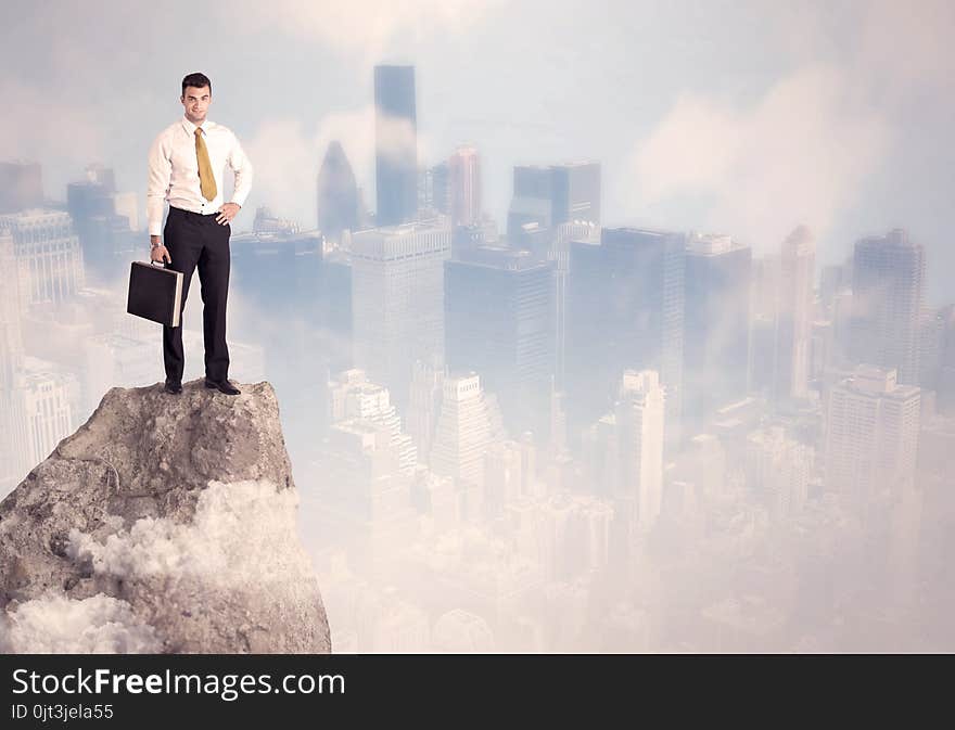 A successful good looking businessman standing on top of a high cliff above the city scape with clouds concept. A successful good looking businessman standing on top of a high cliff above the city scape with clouds concept