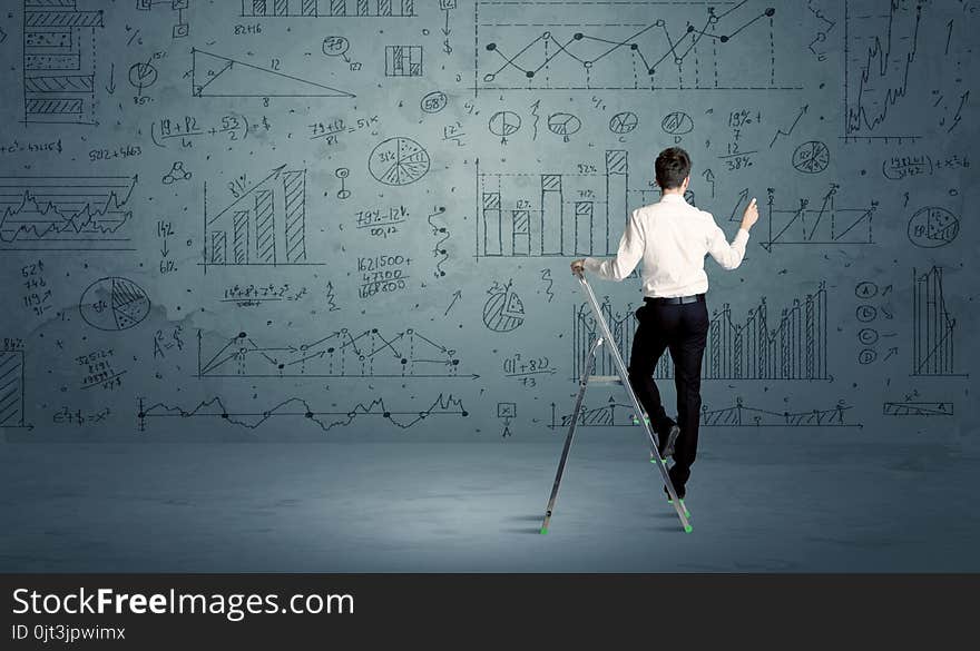 A businessman in modern stylish elegant suit standing on a small ladder and drawing pie and block charts on grey wall background with exponential progressing curves, lines, circles, angles, blocks, numbers. A businessman in modern stylish elegant suit standing on a small ladder and drawing pie and block charts on grey wall background with exponential progressing curves, lines, circles, angles, blocks, numbers