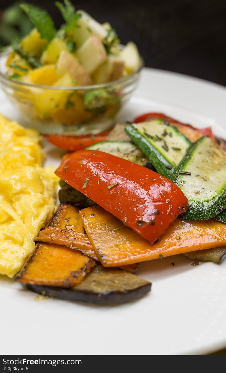 Baked vegetables on white plate: sliced carrots, bell pepper, zucchini with scrambled eggs and blurred mango apple salad in backgr