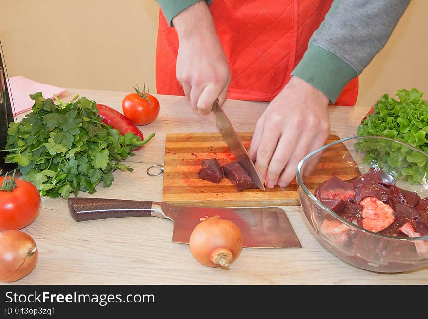 A man in a kitchen apron, meat on a board and a wooden table, a knife and vegetables