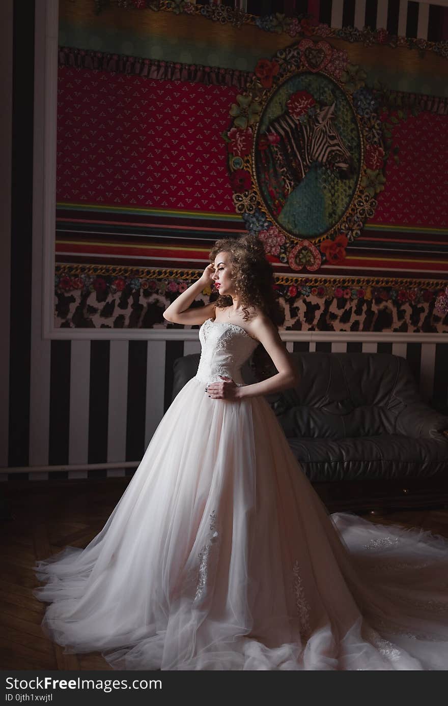 The bride dressed in a chic dress is standing in a room by the window raised her hand to her head. The bride dressed in a chic dress is standing in a room by the window raised her hand to her head